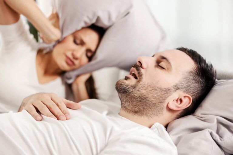 A man snoring and sleeping in the bed while woman is annoyed of it.
