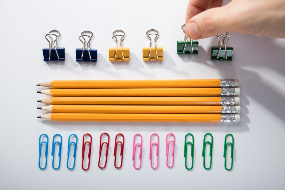 Person's Finger Arranging The Pencils With Row Of Pins Rubber And Pen On White Background