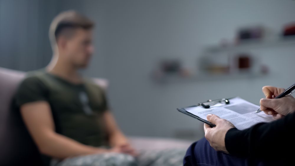 Psychologist making notes during therapy session with sad male soldier