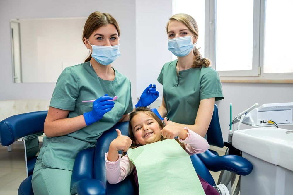 Dentist with assistant and child girl patient showing thumbs up at dental clinic