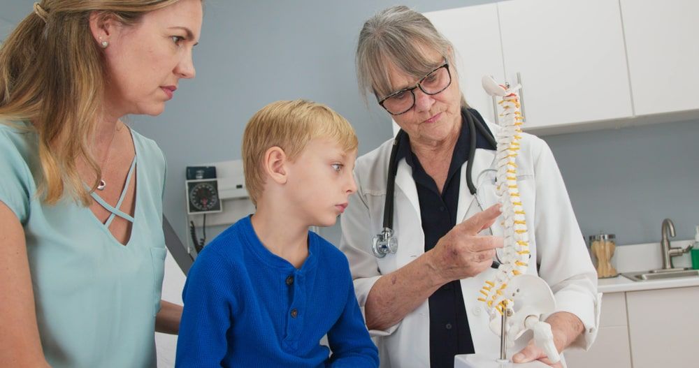 Senior woman pediatrician with spine model talking to little boy patient and his mother about scoliosis