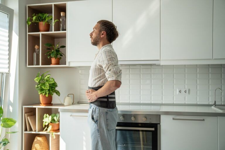 Recovery after spine surgery. Young man wearing corset brace looking out window while standing in kitchen at home