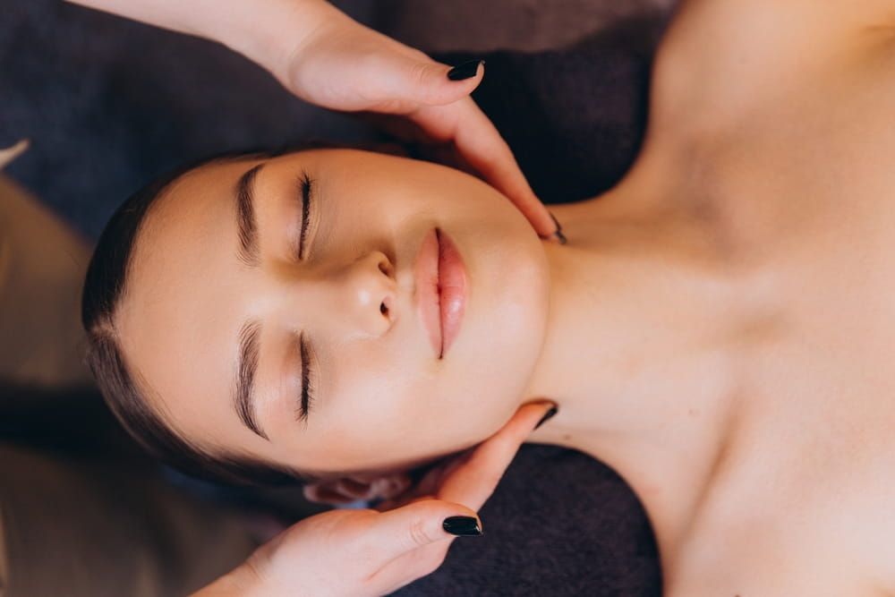 Beautician performing facial massage on young woman lying on massage table with closed eyes and relaxed expression