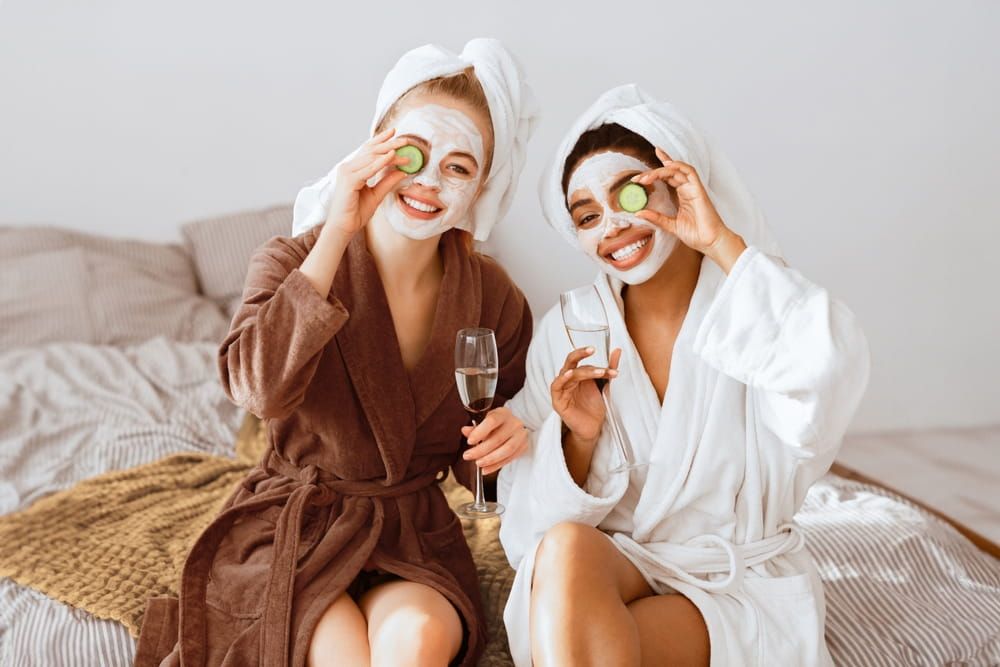 Cheerful multiracial girlfriends with face masks on wearing bathrobes drinking champagne, holding cucumber slices over eyes and smiling at camera, home interior