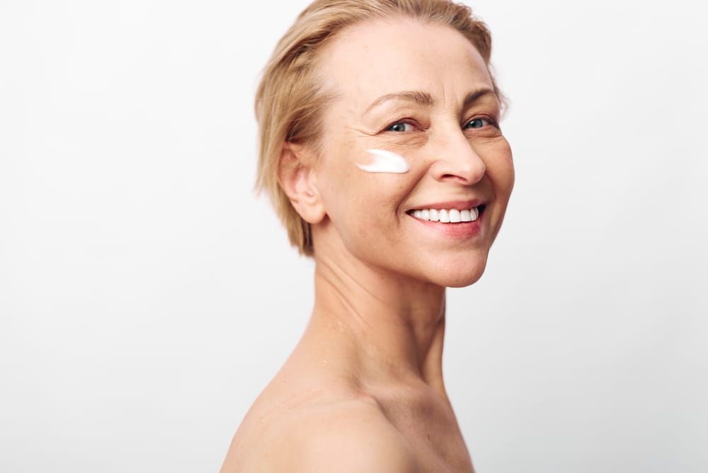 Smiling mature woman with clear skin applies white facial cream on cheek in bright studio.