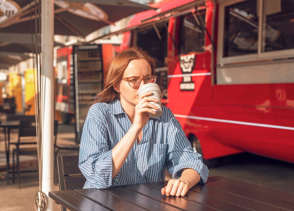 Young woman in eyewear sitting alone with collapsible eco coffee cup near red street food truck