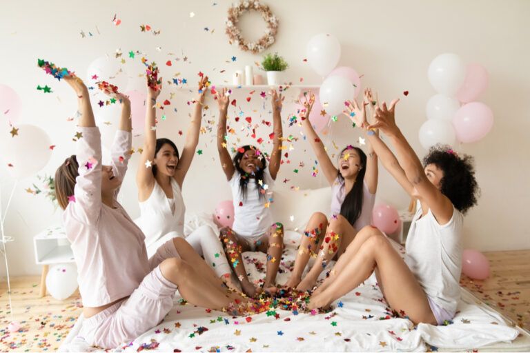 Happy diverse girls sitting on bed in circle, throwing colorful confetti