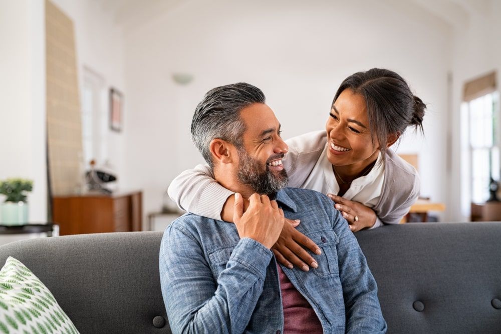 Smiling ethnic woman hugging her husband on the couch from behind