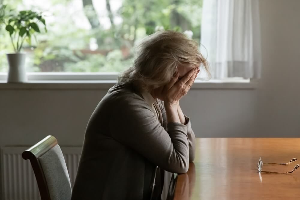 Unhappy old woman sit at table at home