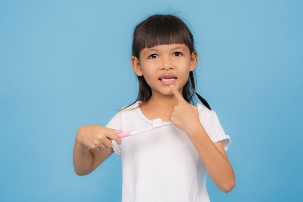 Smiling preschool girl promoting oral hygiene holding a toothbrush isolated on a blue background, teaching children the importance of dental health