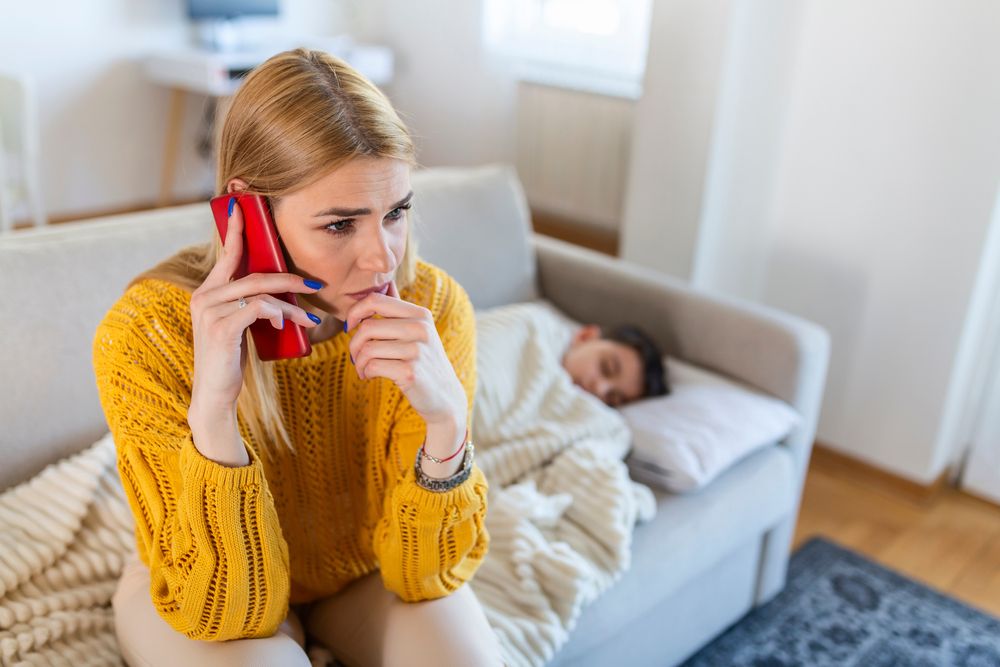 Urgent help. Cheerless troubled mother holding thermometer and calling a doctor while her ill son sleeping