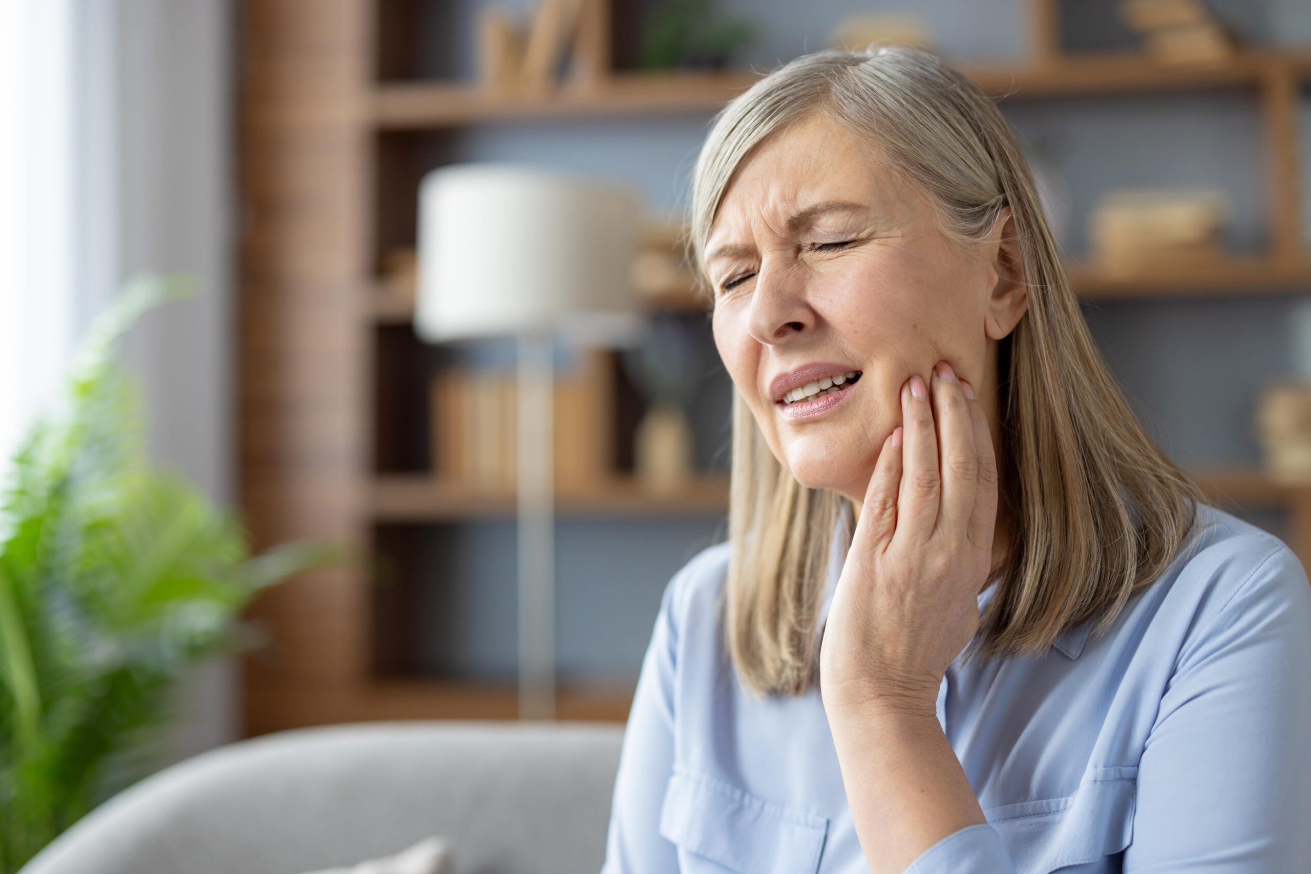 Elderly woman sits at home, touching her cheek and grimacing in pain