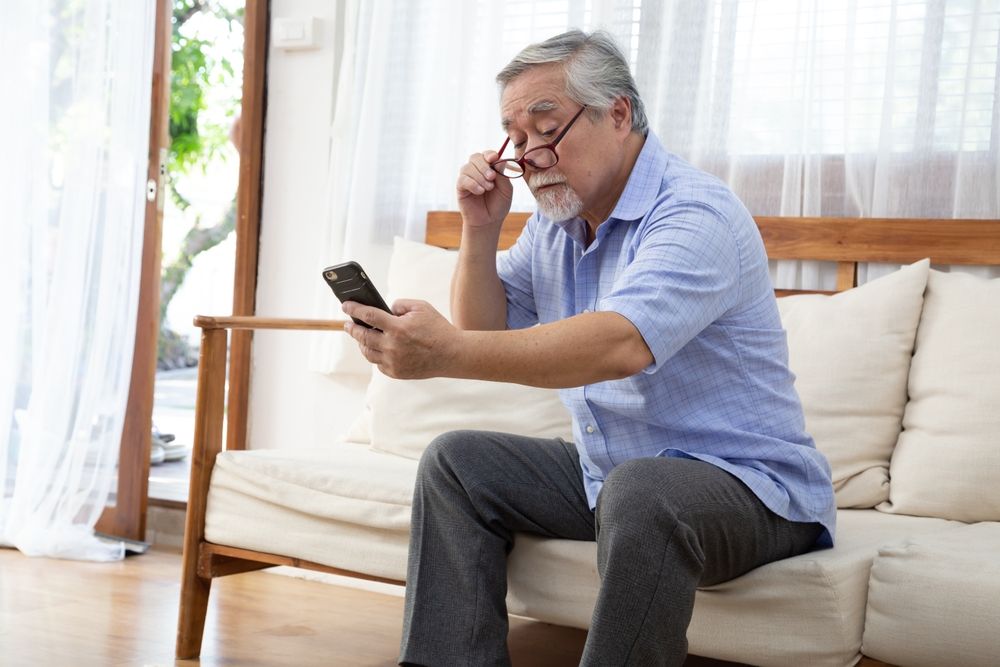 Senior Asian man moves his reading glasses to look at mobile phone while sitting on the living room