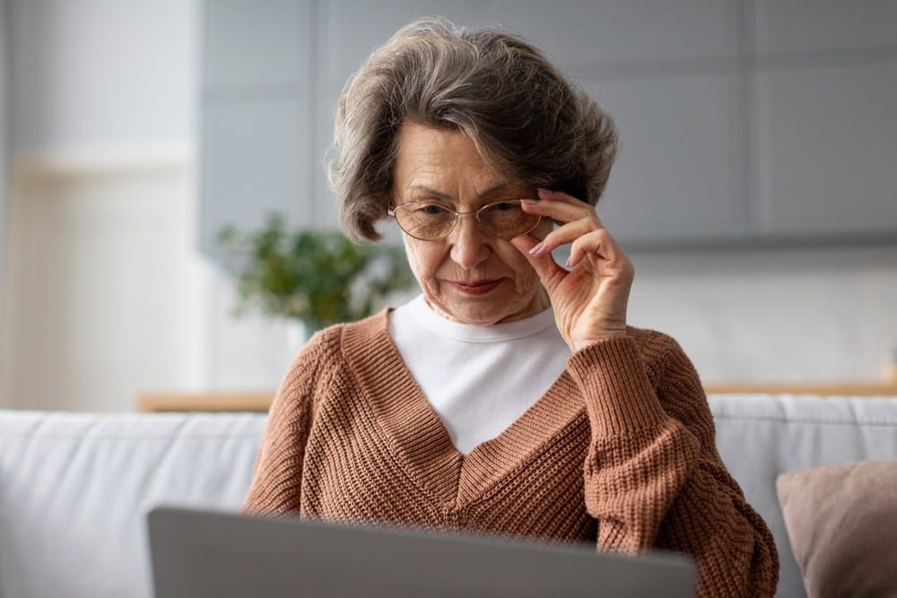 Senior woman using laptop and touching glasses, surfing internet, having problems with vision