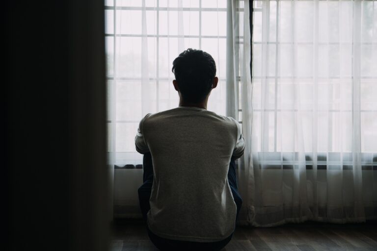 Alone man silhouette staring at the window closed with curtains in bedroom. Man stands at window alone