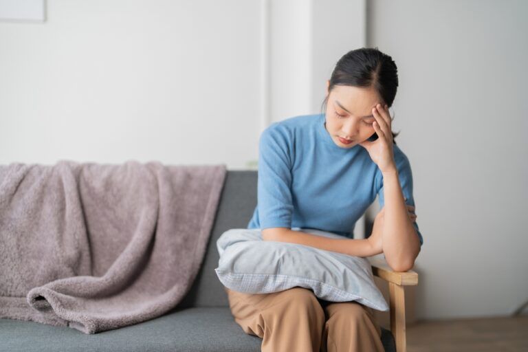 A photograph depicting a female individual in a state of discomfort seated upon a sofa within a residential setting.