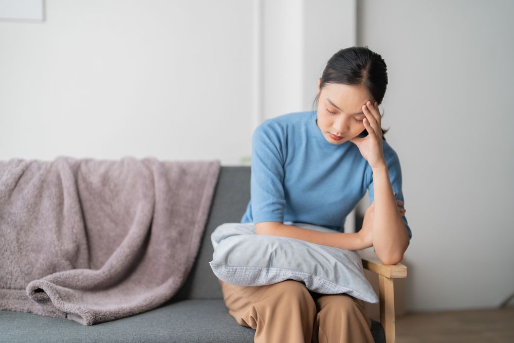 A photograph depicting a female individual in a state of discomfort seated upon a sofa within a residential setting.