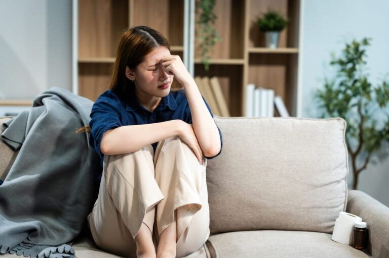 A young Asian woman sits on a sofa in her living room, feeling sick with mental health diseases