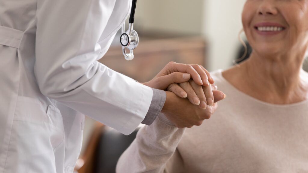 young female physician leaning forward to smiling elderly patient holding her hand