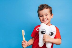 A smiling boy with healthy teeth holds a plush tooth and a toothbrush