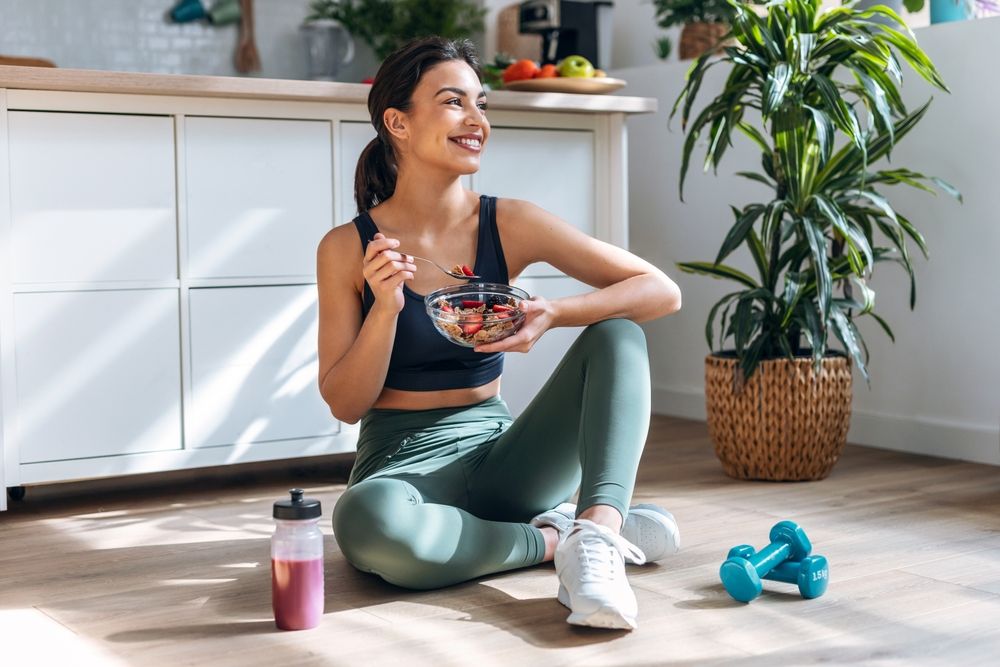 athletic woman eating a healthy bowl of muesli with fruit sitting on floor