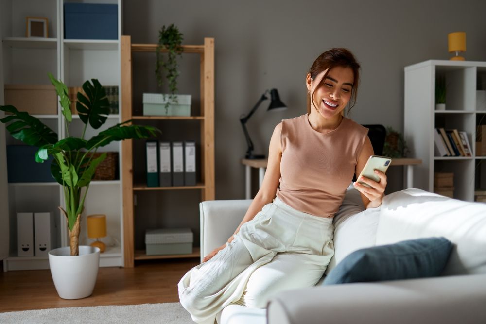 Happy woman using mobile phone and smiling while sitting on sofa at home