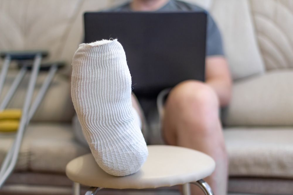 A man sits on a sofa in a cast with a laptop. A leg in a cast lies on a chair