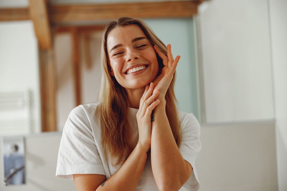 Smiling young woman touching face while standing in bathroom with closed eyes