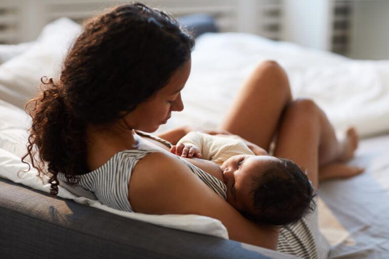 African mother sitting on bed and leaning on headboard while feeding baby with breast
