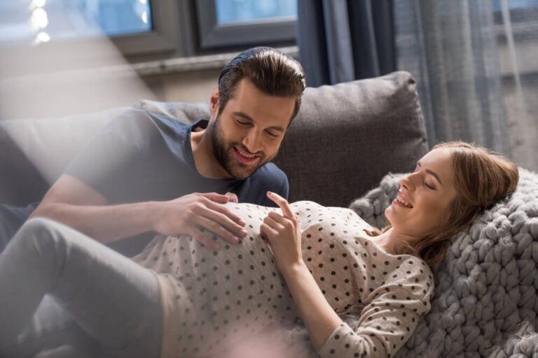 Happy man touching belly of smiling pregnant woman lying on sofa