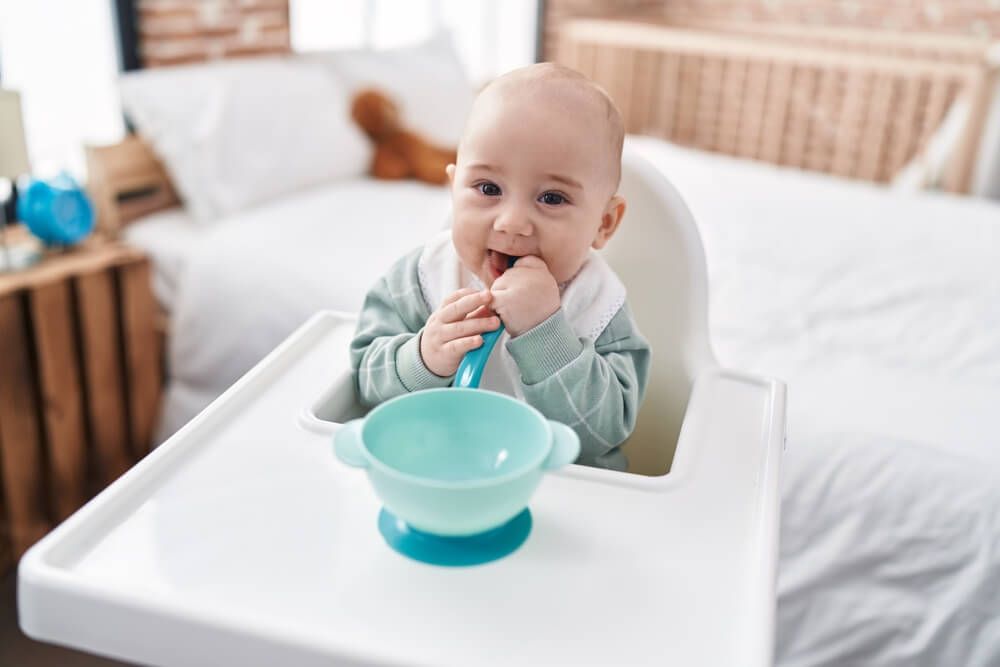 Adorable Caucasian baby sitting on highchair sucking spoon at bedroom