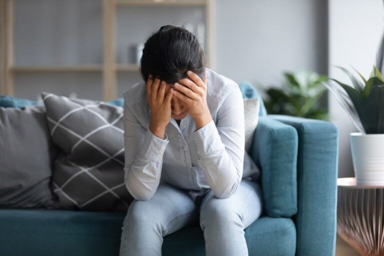 A woman sitting on a couch, looking distressed with her hands on her head.