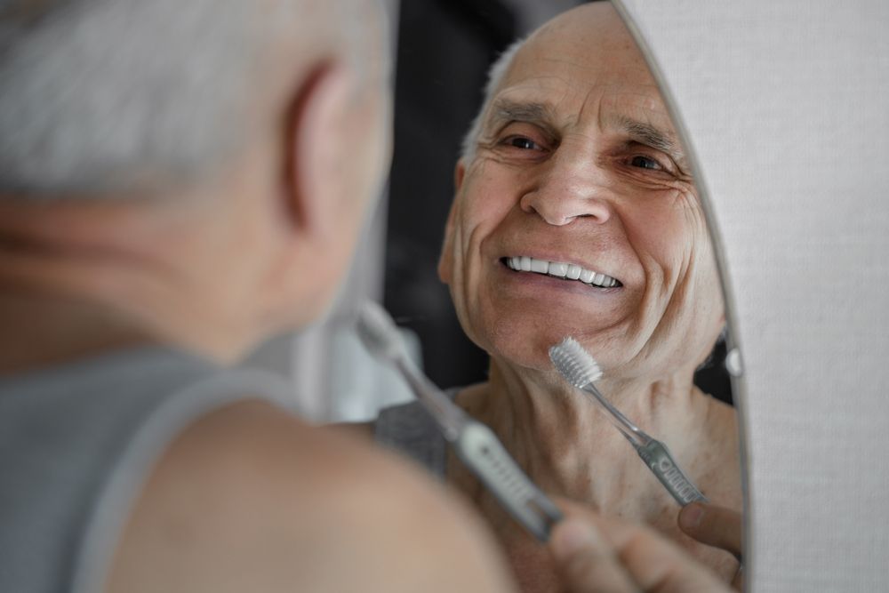 A man smiling in the mirror brushing his dentures