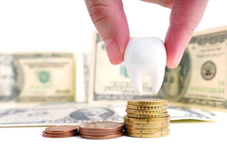 Pinch-holding a large white tooth above coin stacks against dollar bills background