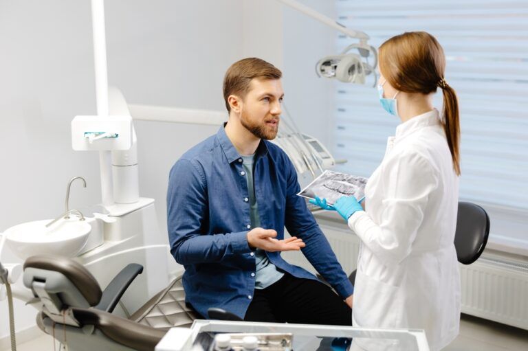 Young attractive man visiting dentist, sitting in dental chair at modern light clinic. Young woman dentist holding x ray image