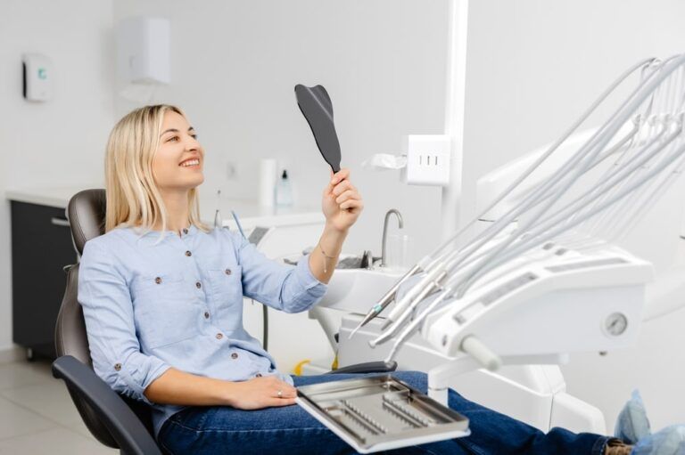 Smiling patient is checking her teeth after dental procedure using small mirror in modern dental clinic
