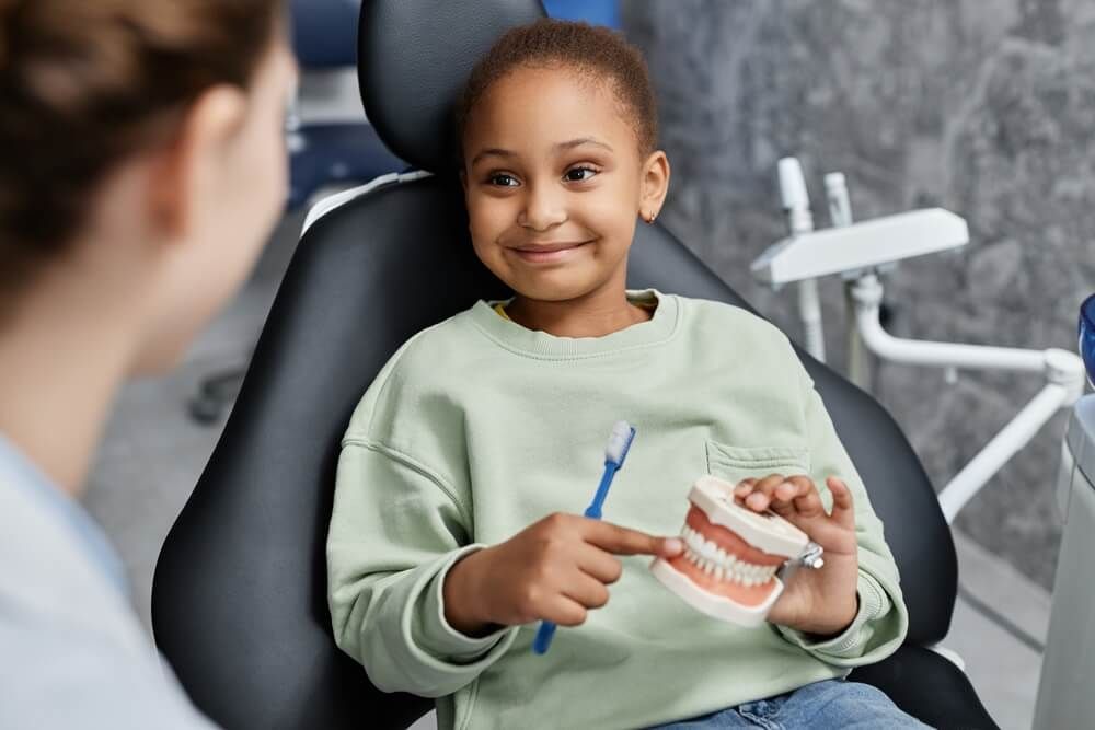 Portrait of black little girl in dental chair holding tooth model and smiling