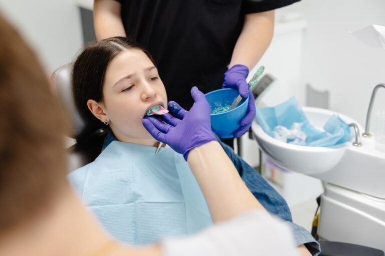 Dentist is using a brush to apply sealant on patient's teeth to prevent cavities, assisted by dental nurse holding saliva ejector